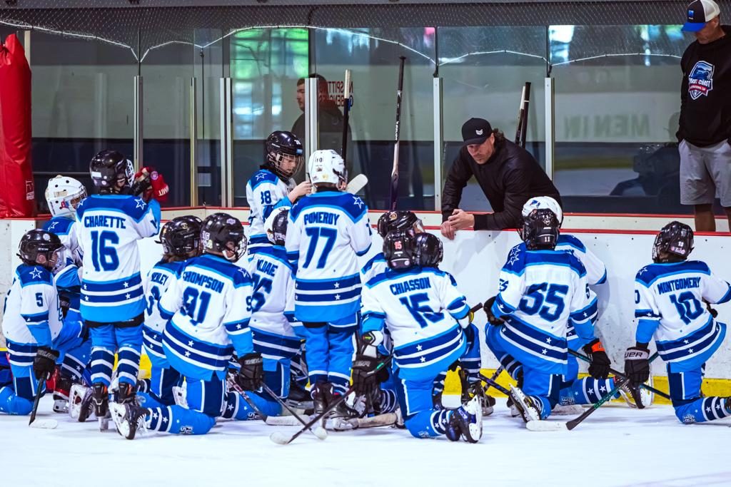 A blue-jerseyed team kneels in a semicircle on the ice while their coach addresses them between shifts