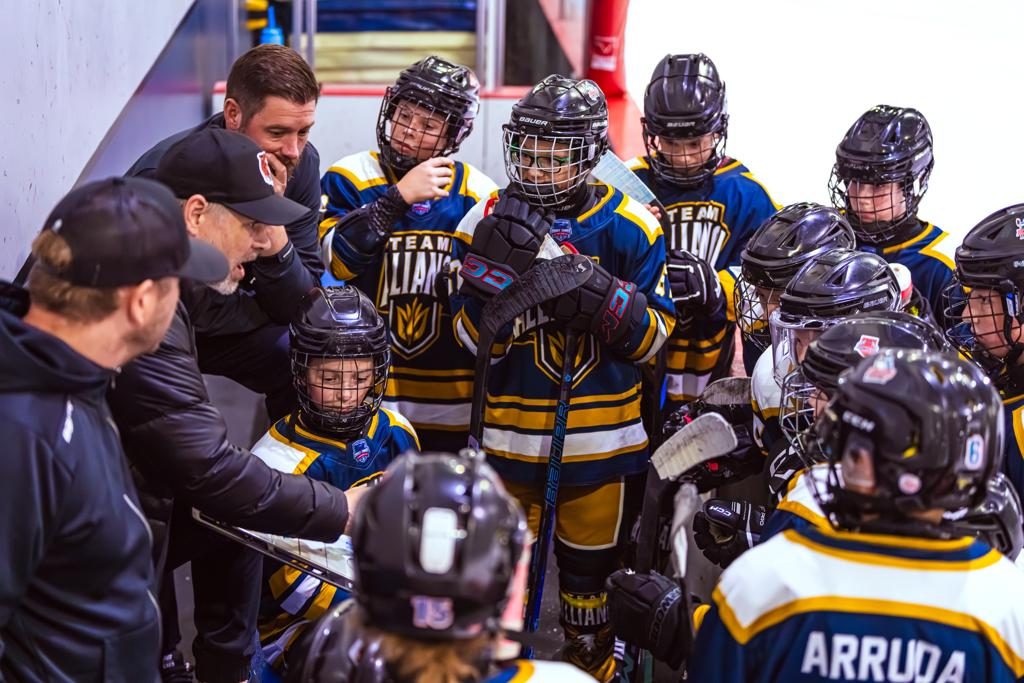 A coach in a suit talks to a huddle of young players in black and gold jerseys gathered around him at the boards