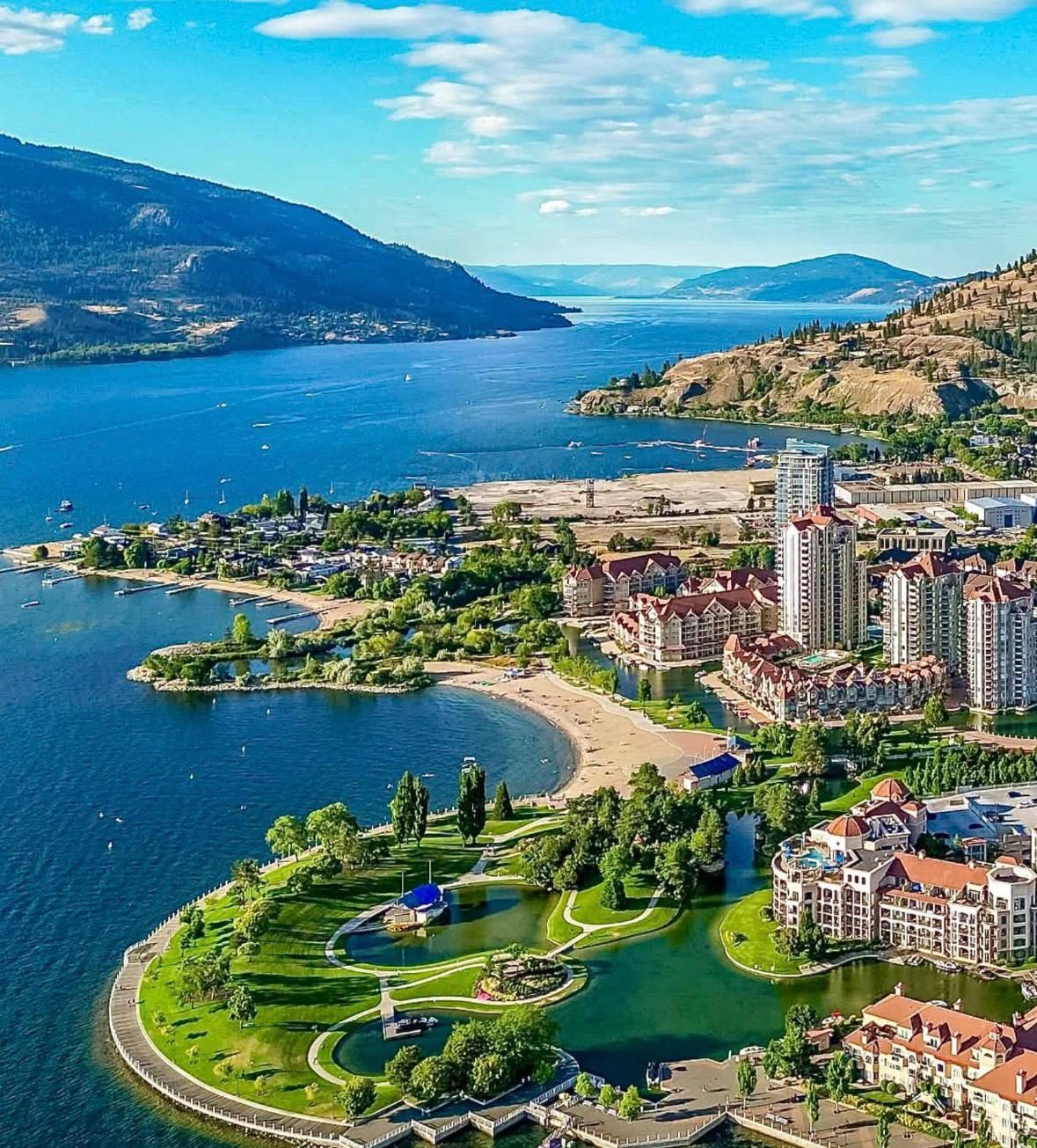 Aerial view of Kelowna's waterfront on Okanagan Lake, with beaches, green parkland and high-rise buildings backed by dry hills