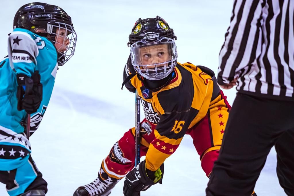 A player in a gold and black jersey battles for the puck against a teal-jerseyed opponent as the referee looks on