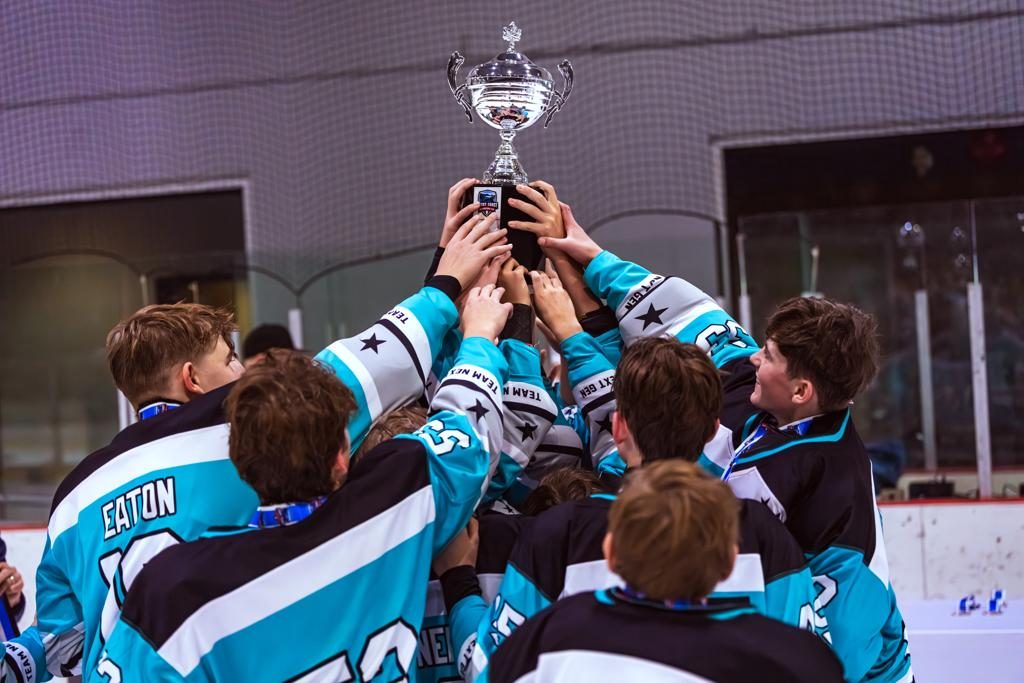 Young players in teal Coast 2 Coast jerseys crowd together to lift the International Cup trophy above their heads on the ice