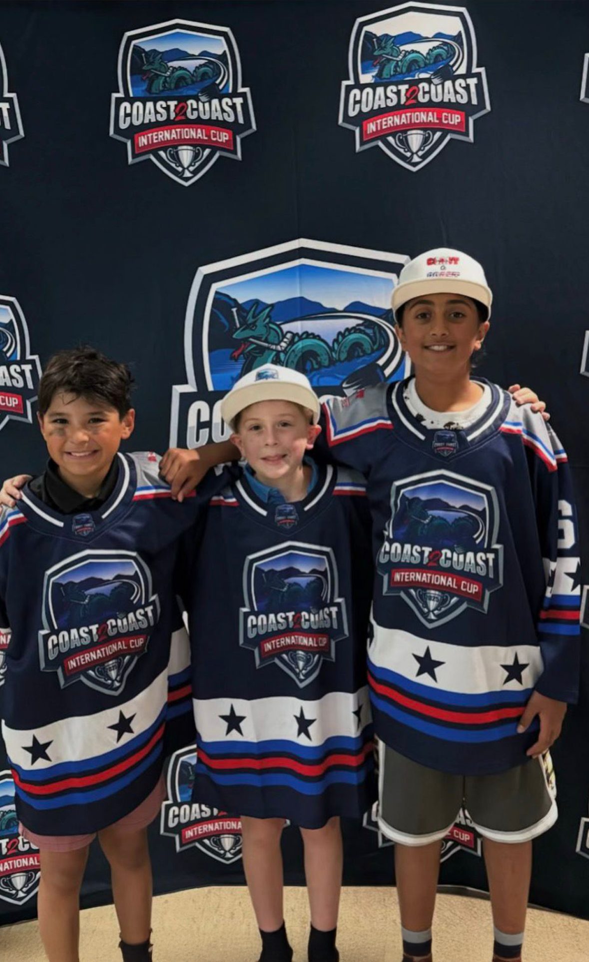 Three young players in navy Coast 2 Coast International Cup jerseys pose smiling in front of the tournament backdrop